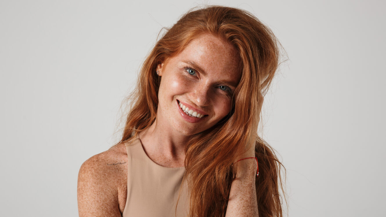 Smiling woman with long red hair and freckled glowing skin against neutral background