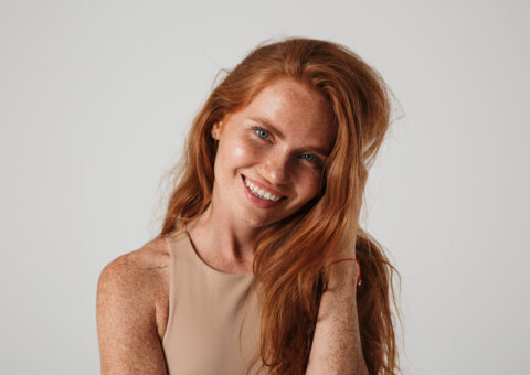 Smiling woman with long red hair and freckled glowing skin against neutral background
