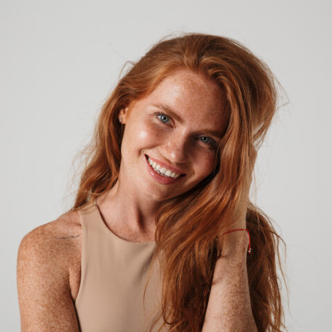Smiling woman with long red hair and freckled glowing skin against neutral background