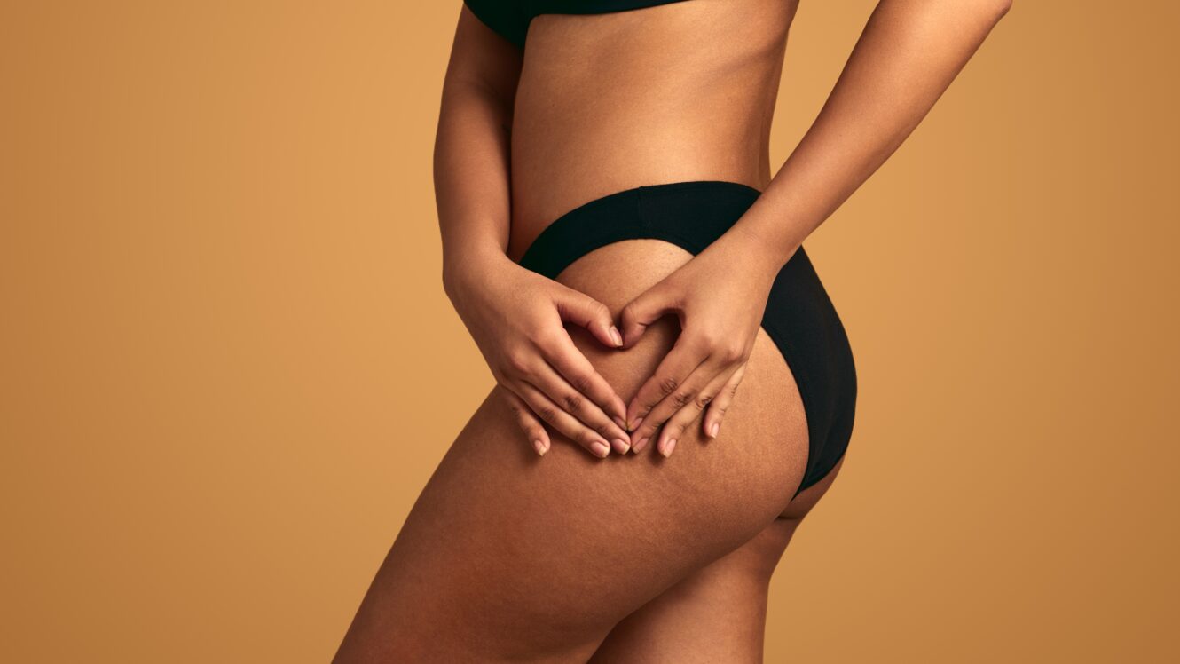 Woman in black underwear shaping her buttocks with hands in a heart symbol, highlighting contour results from butt injections.
