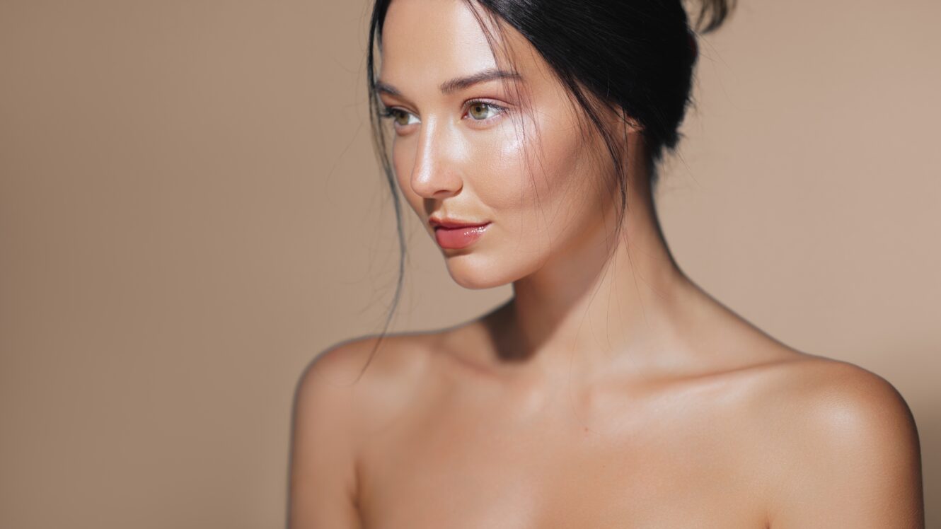 Young woman with glowing, radiant skin and a smooth complexion, looking to the side against a neutral beige background, showcasing natural beauty and skincare results.