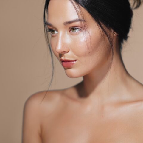 Young woman with glowing, radiant skin and a smooth complexion, looking to the side against a neutral beige background, showcasing natural beauty and skincare results.