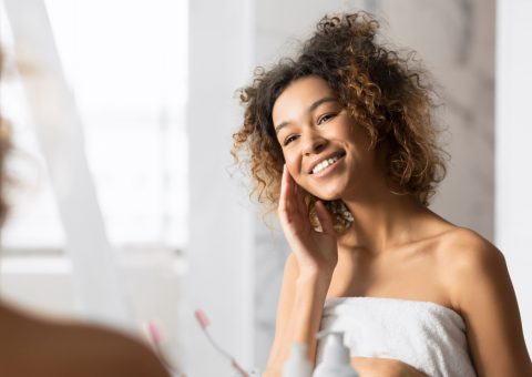 Happy African American Girl Touching Face With Perfect Smooth Skin Looking In Mirror In Bathroom. Selective Focus, Copy Space