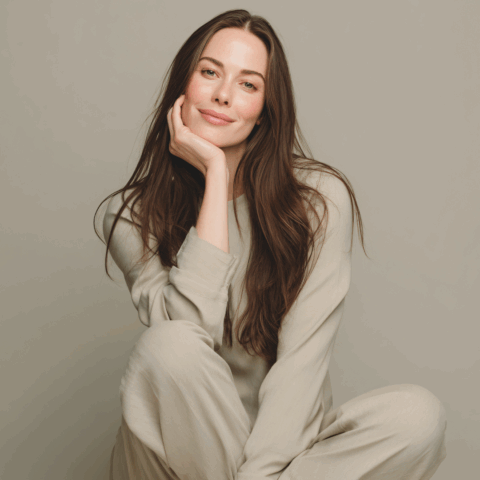 Relaxed woman in neutral loungewear smiling softly with hand on chin against muted background