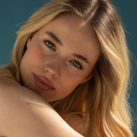 Close-up of a woman with long blonde hair and clear, smooth skin resting her shoulder forward, highlighting natural glow and youthful skin textu