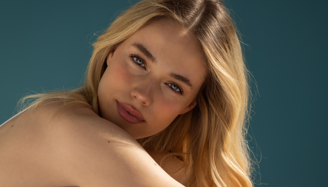Close-up of a woman with long blonde hair and clear, smooth skin resting her shoulder forward, highlighting natural glow and youthful skin textu