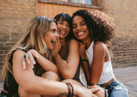 Three women sitting close together outdoors, smiling and laughing with their arms around each other.