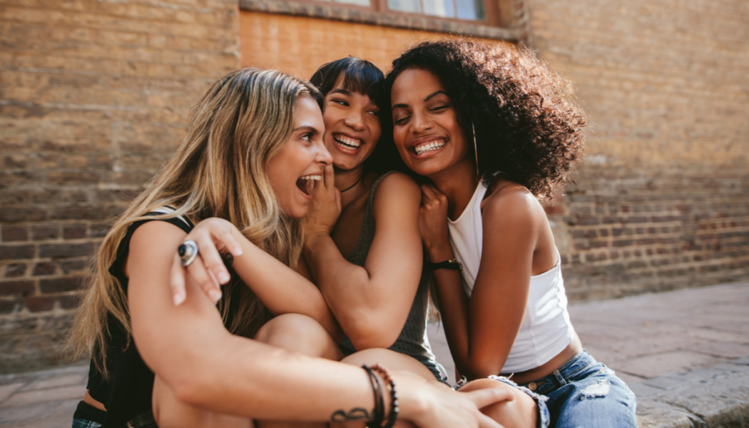 Three women sitting close together outdoors, smiling and laughing with their arms around each other.