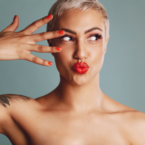 Portrait of a woman with short blonde hair and bright red lipstick posing playfully with her hand near her face, highlighting smooth, youthful skin and facial definition.