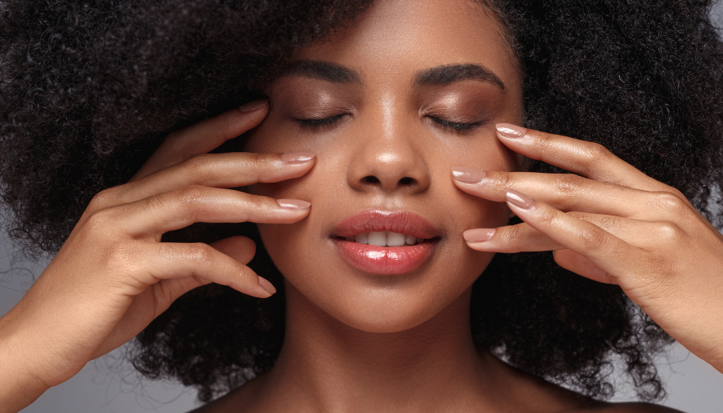 Close-up of a woman with natural curly hair gently touching her face, eyes closed, highlighting smooth skin, softness, and a calm, refreshed appearance.